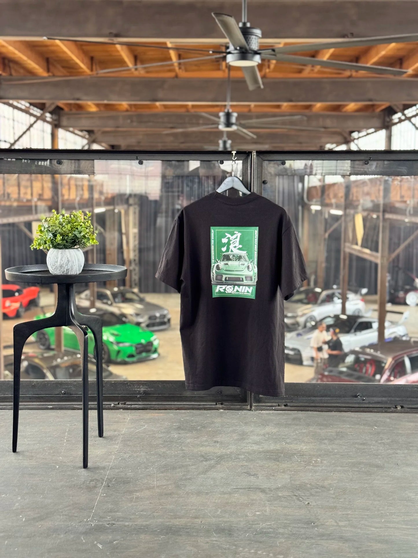 Black t-shirt with a green Porsche GT3RS graphic design hanging in front of a window with a view of cars outside.