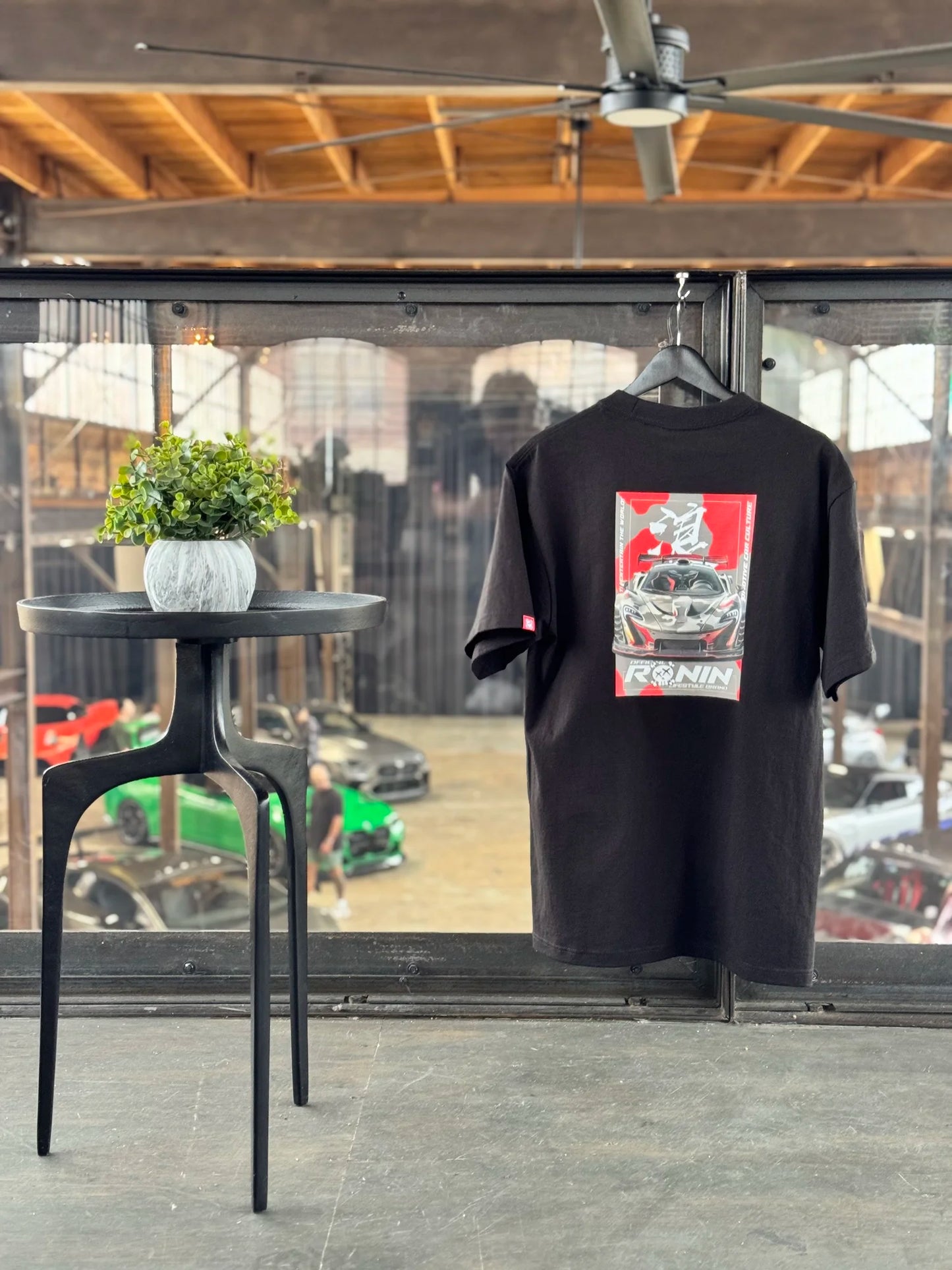 Black t-shirt with a graphic McLaren P1 design on a hanger in a room with a table and plants.