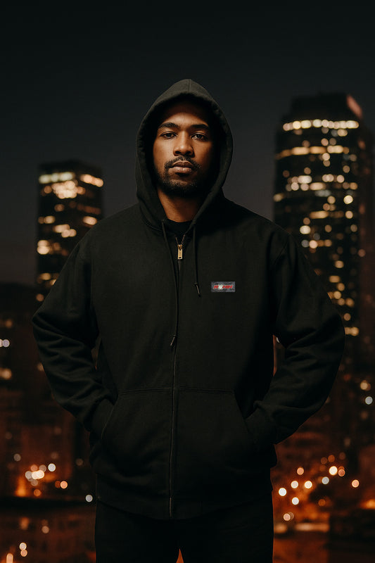 Person wearing a black hoodie with a logo in front of a cityscape at night