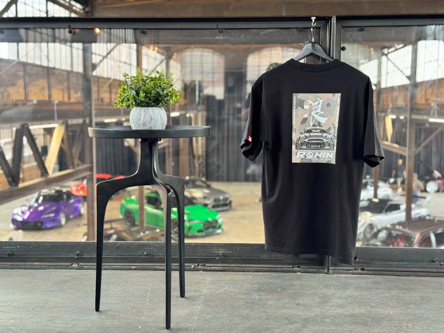 Black t-shirt with a graphic design on a hanger in an industrial setting with cars and a table.