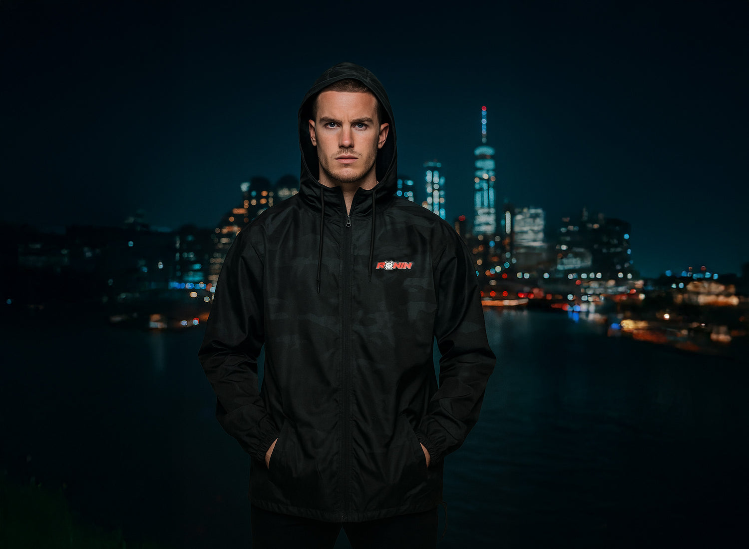 Person wearing a black camo windbreaker jacket with a logo in front of a city skyline at night.